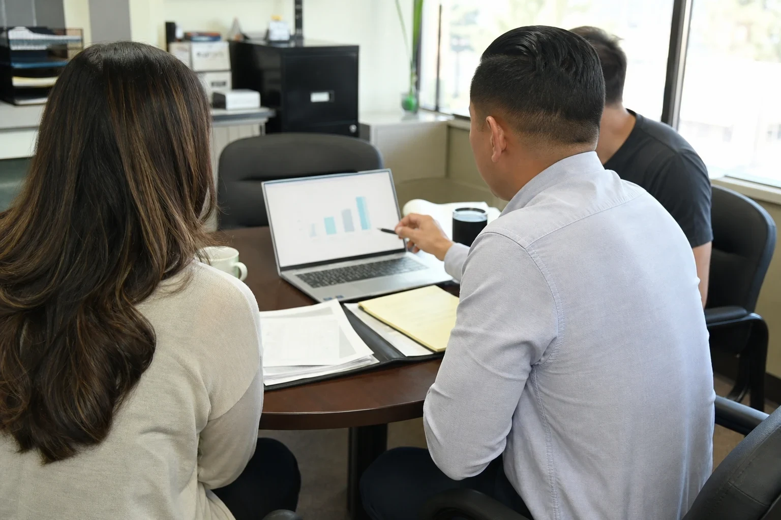 Agent in a light blue button-down points a pen at a bar chart on a laptop while a woman and a man look on at a round office table with papers and a legal pad